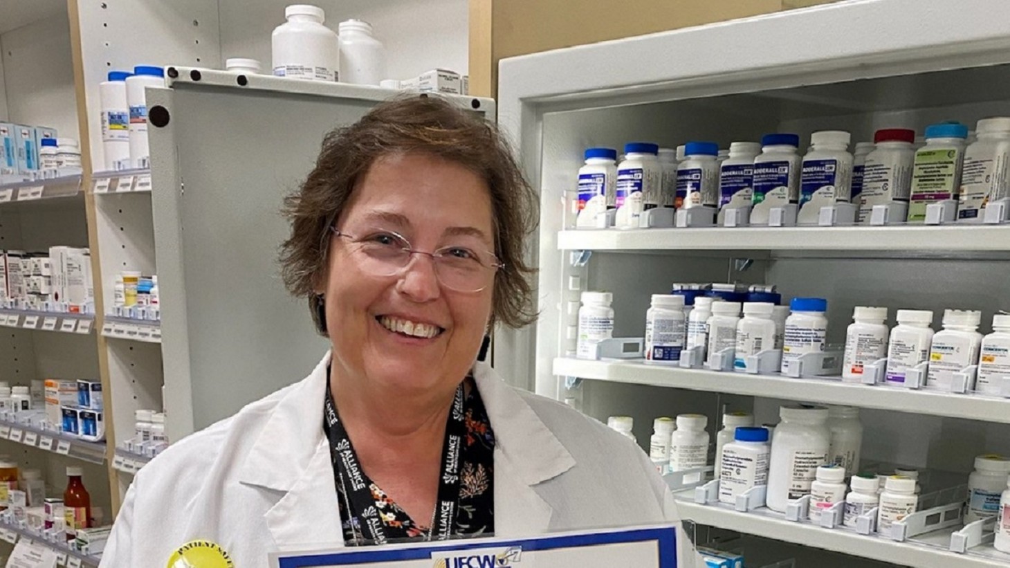 woman in lab coat posing in front of shelves of pill bottles 