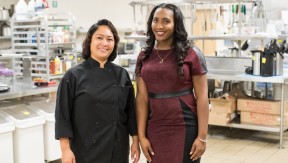 2 women (one wearing a black chef's jacket) in an industrial kitchen
