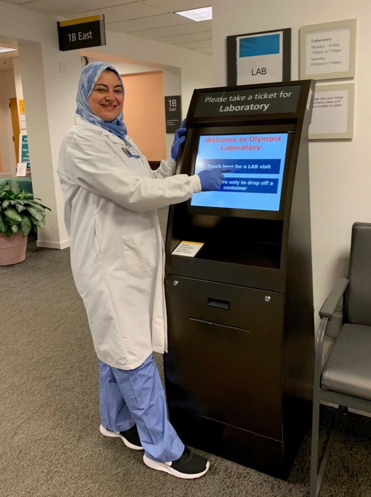 Woman wearing a white lab coat and hijab at a check-in kiosk 