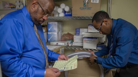 Two men in blue shirts examing packages in a supply room 